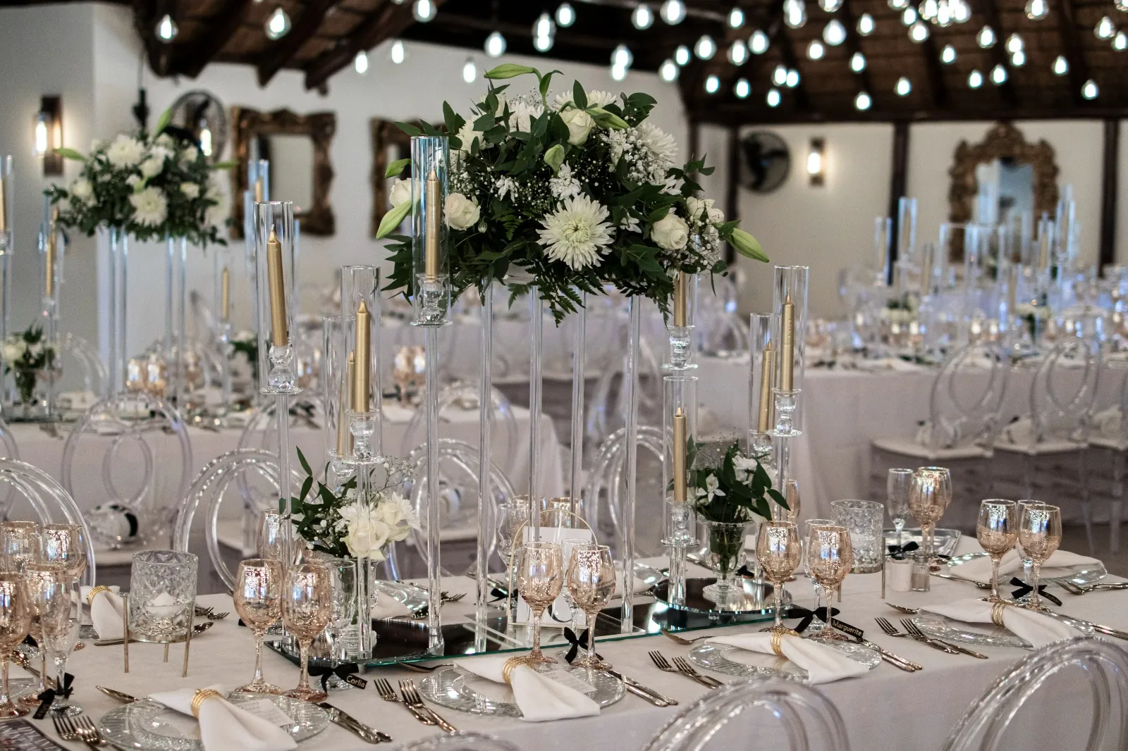 Reception tables with tall white floral centrepieces and gold candles under the festoon lights at Usambara Venue
